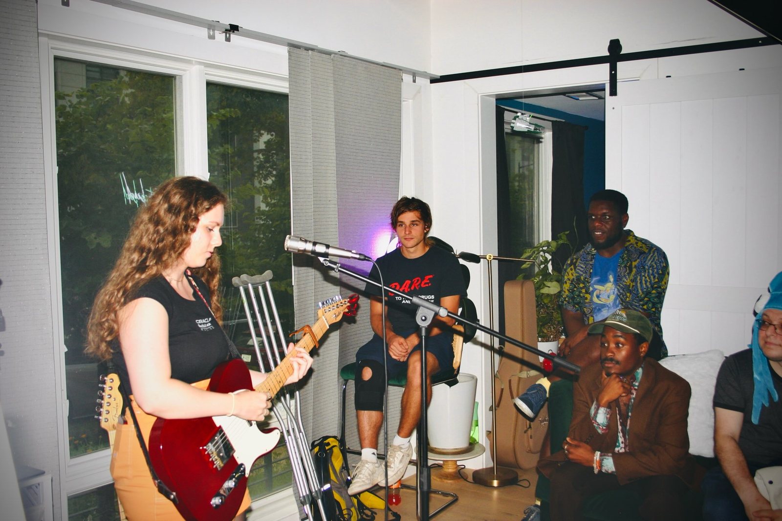 A guitarist with a red electric guitar sings at a microphone while other musicians and the audience look on.