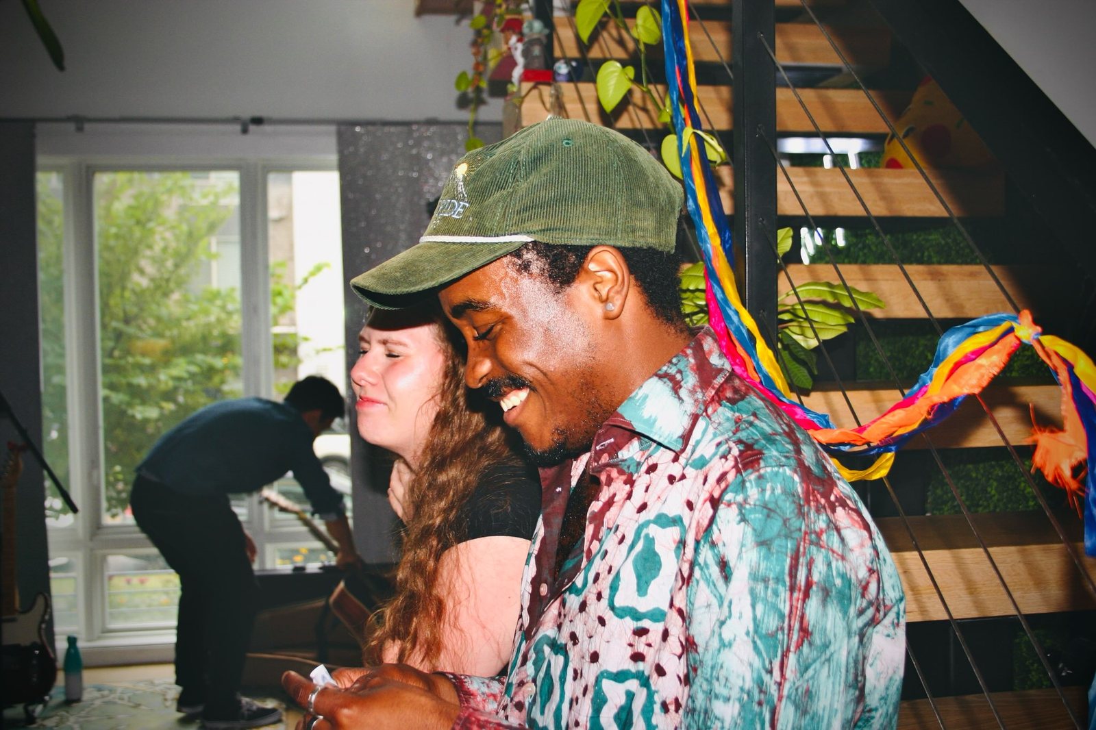 A smiling man in a green cap and patterned shirt beside a staircase hung with colorful streamers.