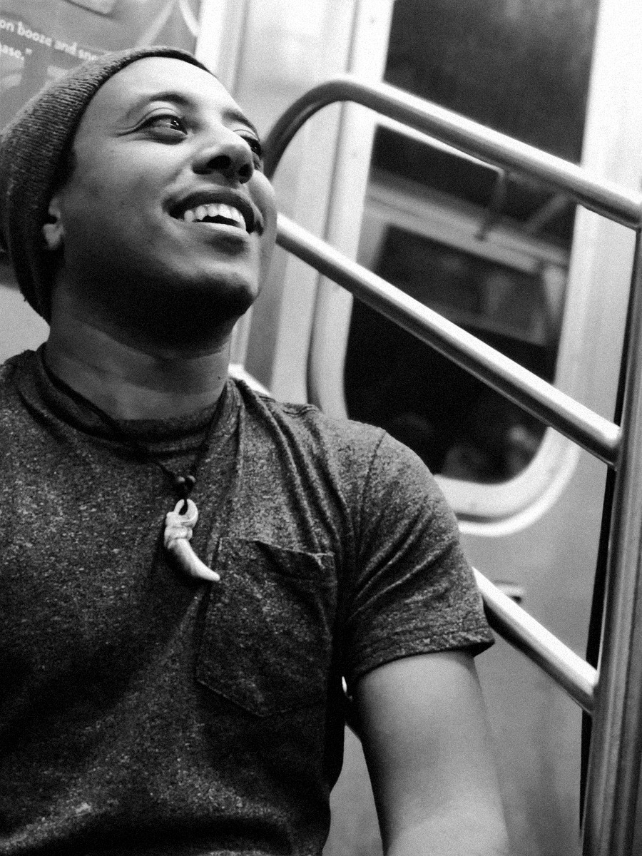 Dag Yeshiwas smiling on a subway train, wearing a knit beanie, a gray t-shirt and a tooth pendant necklace.