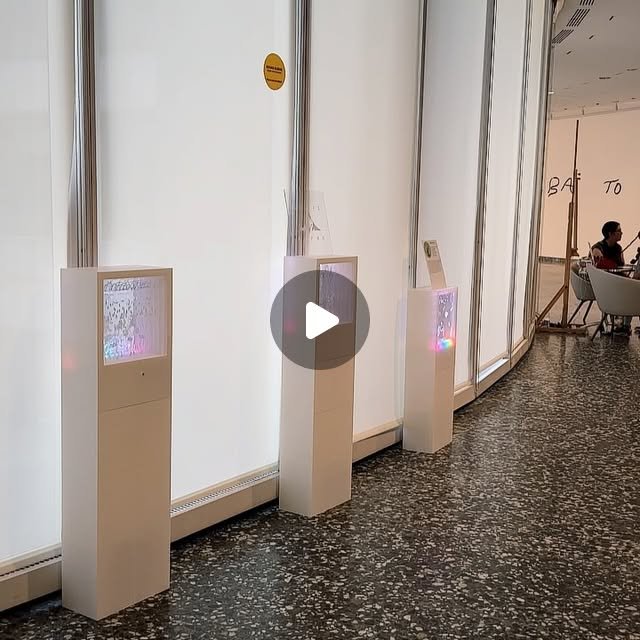 Three white pillars with glowing screens and rainbow lights stand along a curved gallery wall at the Hirshhorn.